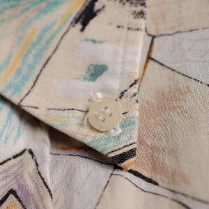 May include: Close-up of a cream-colored button sewn onto a patterned fabric. The fabric features a design with blue, yellow, and black brushstrokes. The button is round with four holes and is attached to the collar of a shirt.