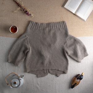 May include: A light brown, off-the-shoulder sweater with short sleeves. The knitwear is displayed on a neutral-toned surface, alongside a cup of tea, a teapot, a book, and a decorative wooden duck.
