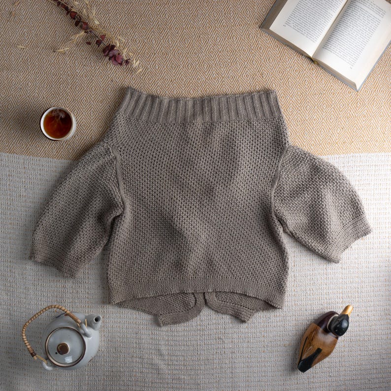 May include: A light brown, off-the-shoulder sweater with short sleeves. The knitwear is displayed on a neutral-toned surface, alongside a cup of tea, a teapot, a book, and a decorative wooden duck.