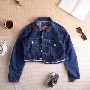 Könnte beinhalten: Dunkelblaue Jeansjacke mit weißem Besatz und rotem, gesticktem Text am Kragen. Die Jacke hat zwei Brusttaschen und eine Knopfleiste. Eine Vintage-Kamera und Sonnenbrille sind ebenfalls im Bild.