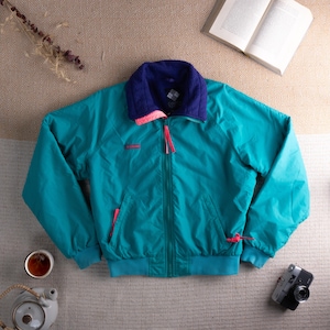 Könnte beinhalten: Türkis- und marineblaue Jacke mit pinkfarbenem Kragen und Reißverschlüssen. Die Jacke hat einen durchgehenden Reißverschluss und zwei Vordertaschen. Das Columbia-Logo ist sichtbar. Vintage-Kleidungsstück, ideal für Outdoor-Aktivitäten. Maße in cm.