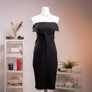 Könnte beinhalten: Schwarzes trägerloses Kleid auf einer Schaufensterpuppe. Das Kleid hat eine Bindedetail an der Taille und eine Midi-Länge. Eine silberne Halskette ist zu sehen. Der Hintergrund umfasst ein weißes Regal mit Büchern und Dekorationsartikeln.