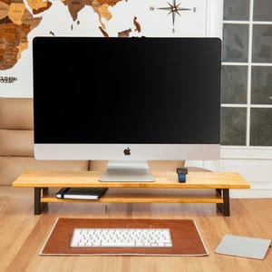 May include: A wooden monitor stand with a black metal frame supports a silver iMac computer. A keyboard, mousepad, and a small notebook are on the desk. A world map and a compass rose are on the wall.