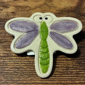 Puede incluir: Un broche de cerámica con forma de libélula, con cuerpo crema, alas moradas y abdomen verde. La libélula tiene ojos negros y está sobre un fondo de madera. Este broche decorativo es un accesorio caprichoso.