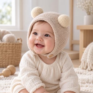 May include: A beige textured knit bonnet featuring two round pom-poms on top and a small wooden button closure under the chin. The hat is crafted from a soft, fuzzy material and provides full coverage for the head, designed with a snug fit.