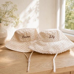 以下が含まれることがあります： 「Emma」と「Lily」と刺繍された、ベージュのドット柄バケットハット2つ。片方の帽子にはレースのトリムが付いています。どちらにもあご紐が付いています。木製の表面に置かれ、背景には窓があります。