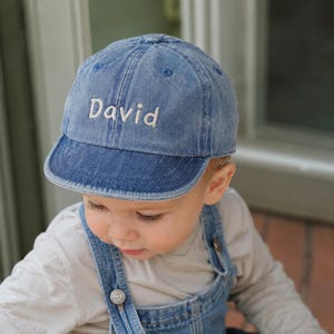 May include: A denim baseball cap with the name "David" embroidered in white. The cap is a light blue color and has a curved brim. The cap is worn by a child.