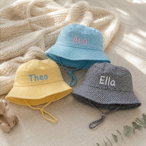 May include: Three bucket hats in blue, yellow, and black gingham patterns. Each hat features an embroidered name on the front: Ava in pink, Theo in blue, and Ella in white. Each hat includes a matching chin strap with an adjustable toggle.