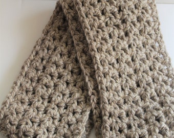 May include: A folded, chunky-knit scarf in a neutral beige color. The scarf is made of thick yarn, with a textured, open weave pattern. The scarf is rectangular and appears soft and warm, suitable for cold weather.