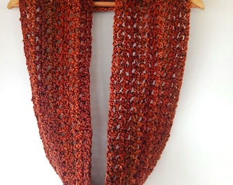 May include: A chunky crochet infinity scarf in a variegated blend of deep rust, burnt orange, and dark brown yarn. The accessory features an open, airy stitch pattern and is displayed hanging on a wooden clothes hanger.