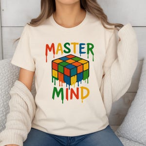 May include: A cream-coloured t-shirt with a colourful Rubik's Cube design and the words "MASTER MIND" in a dripping paint style. The design uses red, orange, yellow, green, blue, and black. The shirt has a crew neck and short sleeves.