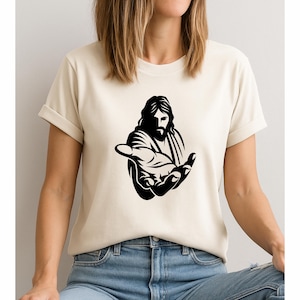 Puede incluir: Camiseta beige con un gráfico negro de una figura religiosa extendiendo una mano. El diseño representa una figura con pelo largo y barba, ofreciendo un gesto de bienvenida. La camiseta es de material suave y tiene mangas enrolladas.