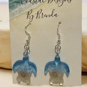 May include: A pair of earrings featuring a sea turtle design. The earrings are made of clear resin with blue and white accents, and a silver-colored hook. The card behind the earrings reads "Coastal Designs By Brenda".