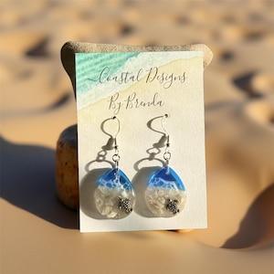 May include: Teardrop-shaped earrings with a coastal theme. The earrings feature a blue and white resin design, with small turtle charms and shell accents. The card behind the earrings reads "Coastal Designs By Brenda."