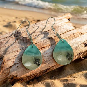 May include: Teardrop-shaped earrings with a beach theme. The top half is turquoise, the bottom half is sandy, and a small silver turtle charm is attached. The earrings hang from silver hooks and are displayed on a piece of driftwood.