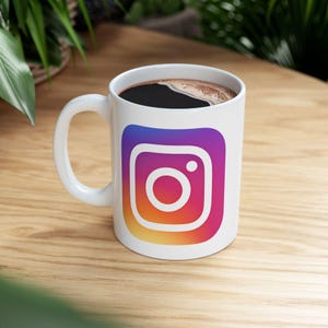 May include: White ceramic mug with a colorful Instagram logo printed on the front. The mug is filled with coffee.