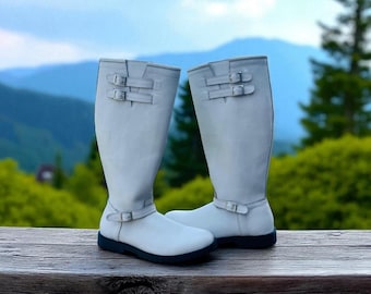 May include: A pair of light gray knee-high boots with black soles. Each boot features two decorative buckles at the top and one at the ankle. The boots are made of a smooth material and are set against a blurred outdoor background.