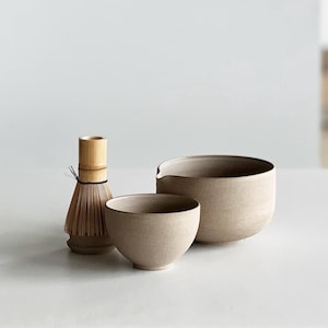 May include: A matcha tea set comprising a bamboo whisk, a small bowl, and a larger bowl with a spout. The items are a neutral beige colour and appear to be ceramic. The set is arranged on a white surface.