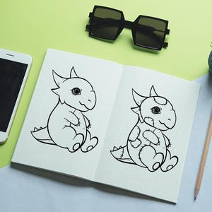 May include: Open notebook with two cartoon dragon illustrations. One dragon is plain, the other has patches. A smartphone, sunglasses, a small plant, and a pencil are also on the light green and blue surface.