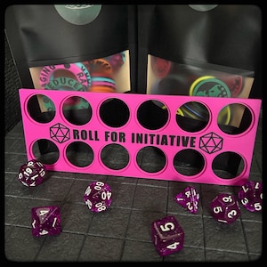 May include: A bright pink dice holder with the text "ROLL FOR INITIATIVE" and a D20 symbol. Several purple dice are scattered around the holder. Two black bags with colorful game pieces are in the background.
