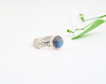 Puede incluir: Un anillo de plata con una banda texturizada y una piedra redonda de labradorita azul grisácea.
