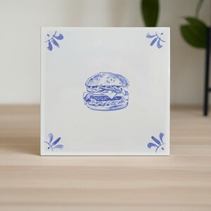 Op de afbeelding: Vierkante witte keramische tegel met een blauwe illustratie van een hamburger. De tegel heeft blauwe bloemenhoekaccenten. De burgerillustratie bevat de broodjes, patty, kaas en andere toppings.