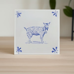 May include: White square tile with a blue illustration of a goat. The goat has a bell around its neck. Blue floral accents are in each corner. The tile is on a wooden surface.