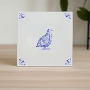 Könnte beinhalten: Quadratische weiße Keramikfliese mit einer blauen Illustration eines Vogels. Der Vogel ist mittig auf der Fliese platziert, mit blauen floralen Eckakzenten. Die Fliese wird auf einer hellen Holzoberfläche präsentiert.
