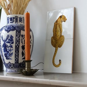 May include: A ceramic tile with a leopard illustration in yellow and brown spots on a white background. The tile is displayed on a marble surface, next to a blue and white vase and a lit orange candle in a brass holder. The candle is orange.