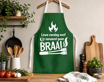 Op de afbeelding: Een donkergroen schort met de witte tekst "Lewe vandag asof jy vanaand gaan BRAAI", een vlamicoon en een afbeelding van een barbecuetang. Het schort heeft witte nek- en taillebanden en hangt aan een houten kledinghanger.