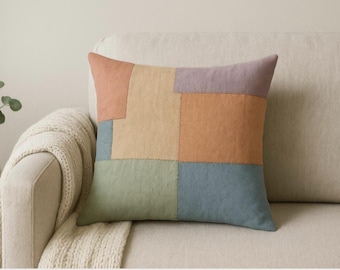 Puede incluir: Un cojín cuadrado con un diseño de patchwork compuesto por paneles de tela rectangulares en tonos apagados de melocotón, lavanda, amarillo mostaza, verde salvia y azul pizarra, unidos con costuras visibles.