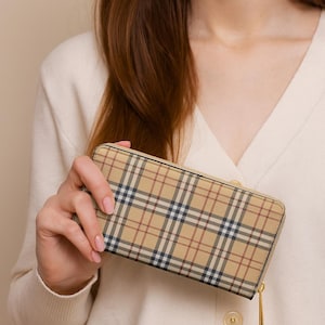 May include: A beige wallet with a classic plaid pattern in tan, navy, and red. The wallet has a gold-colored zipper and is held by a person wearing a cream-colored cardigan. The wallet is a rectangular shape.