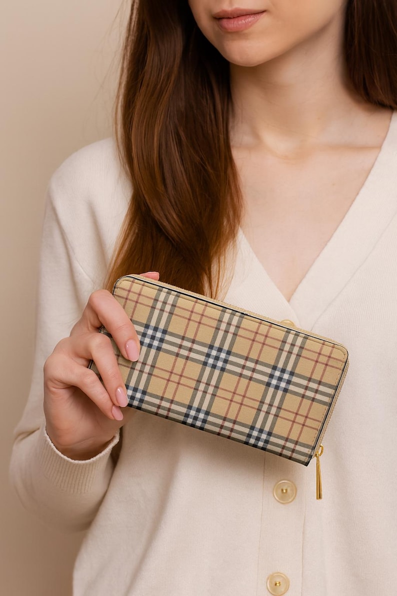 May include: A beige wallet with a classic plaid pattern in tan, navy, and red. The wallet has a gold-colored zipper and is held by a person wearing a cream-colored cardigan. The wallet is a rectangular shape.