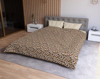 Puede incluir: Una funda nórdica con un patrón repetitivo de leopardo en tonos beige, marrón, negro y rosa suave. La ropa de cama cubre una cama con un cabecero gris capitoné y dos almohadas rectangulares de color crema colocadas en la parte superior.
