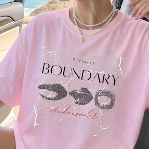 Könnte beinhalten: Hellrosa T-Shirt mit dem Wort "BOUNDARY" in Schwarz und dem Wort "Ambassador" in Schreibschrift. Das Shirt hat eine Grafik von drei dunkelgrauen Formen. Das Wort "Official" steht über "BOUNDARY". Das Shirt ist unten mit "SOFTPINK" beschriftet.