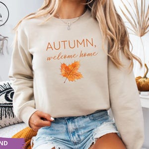 Puede incluir: Una sudadera de cuello redondo color arena que presenta el texto "AUTUMN, welcome home" en letras naranjas y marrones, acompañado de una ilustración estilo acuarela de una sola hoja de arce naranja centrada en la parte frontal.
