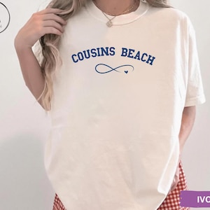 May include: Ivory-colored t-shirt with the text "COUSINS BEACH" in navy blue, featuring an infinity symbol and a small heart. The shirt is a relaxed fit, suitable for casual wear.