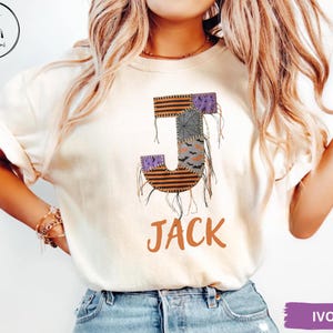 Puede incluir: Una camiseta color marfil con una letra J grande diseñada con un patrón de retazos de rayas naranjas, murciélagos morados y telarañas grises, con hilos deshilachados. El nombre JACK está impreso debajo en letras naranjas sin remates.