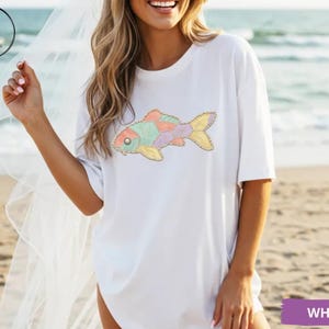 May include: White t-shirt featuring a colorful fish design. The fish has a mix of pastel colors including pink, green, yellow, and purple. The shirt is a casual, oversized style.