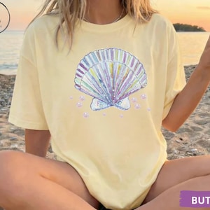 Puede incluir: Camiseta de color mantequilla con un diseño de concha en colores pastel y detalles de perlas. La camiseta tiene cuello redondo y mangas cortas. La palabra "BUTTER" está impresa en un rectángulo morado en la parte inferior derecha.