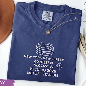 May include: Navy blue t-shirt with a white graphic of a stadium and text. The text includes the location, coordinates, and date: "NEW YORK NEW JERSEY 40.8135° N 74.0745° W 19 JULIO 2026 METLIFE STADIUM". A gold necklace is draped over the shirt.
