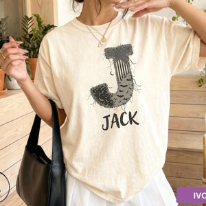 May include: An ivory short-sleeve t-shirt featuring a large letter J graphic with a distressed, textured pattern containing bats and stripes. Below the letter, the name JACK is printed in a bold, black, sans-serif font. The shirt has a crew neckline.