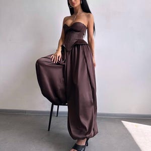 May include: A dark brown strapless corset top with a structured bodice and matching high-waisted, wide-leg satin trousers that feature a voluminous, gathered silhouette. The outfit is paired with simple black open-toe heeled sandals.