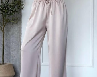 May include: High-waisted, wide-leg trousers in a pale champagne satin fabric. The trousers feature an elasticated waistband with a matching drawstring tie at the centre front, creating a relaxed, flowing silhouette that falls to the floor.