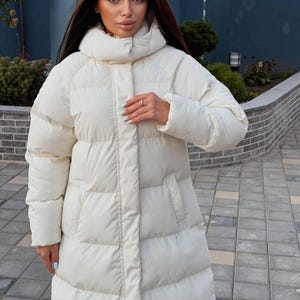 Può includere: Un lungo piumino color bianco sporco caratterizzato da un colletto alto, impunture orizzontali e una chiusura centrale con zip nascosta da una patta. Il cappotto presenta maniche lunghe e due tasche laterali, offrendo una silhouette strutturata.