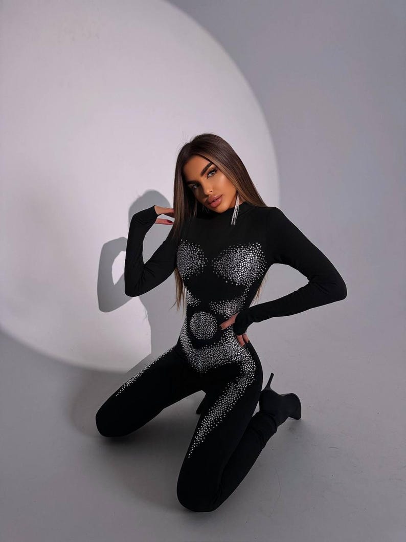 May include: Black jumpsuit with long sleeves and a high neckline. The jumpsuit is embellished with sparkling rhinestones in a decorative pattern across the chest, waist, and legs. The model is wearing black high-heeled boots.
