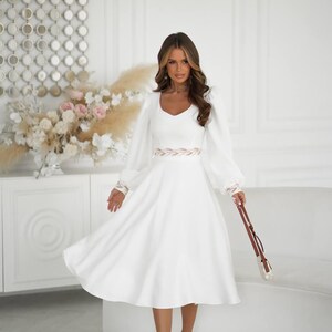 May include: A white dress with long, puffy sleeves and a V-neckline. The dress features a decorative belt and is paired with brown heels and a small purse. The background includes a white sofa and decorative wall art.