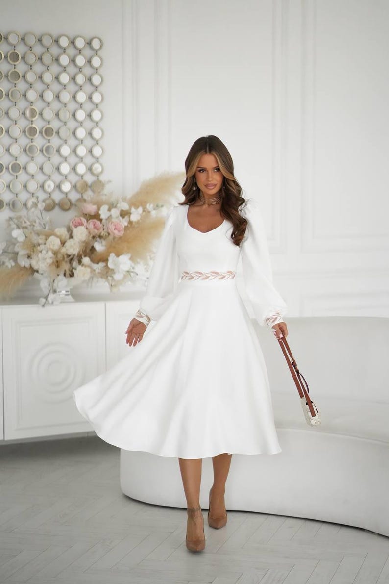 May include: A white dress with long, puffy sleeves and a V-neckline. The dress features a decorative belt and is paired with brown heels and a small purse. The background includes a white sofa and decorative wall art.