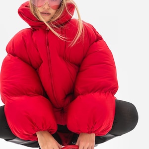 Peut inclure: Une doudoune rouge vif avec une silhouette oversize, un col montant et des manches longues à poignets élastiqués. La veste présente une fermeture éclair verticale centrale et un tissu à la finition mate sur tout le vêtement.