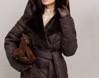 May include: A dark brown quilted puffer coat featuring a large hood with a dark brown faux fur trim, a matching tie belt at the waist, and double-breasted button closures. The coat is worn over a beige mock neck sweater and paired with brown gloves.